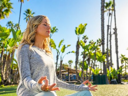 A woman meditates on a sunny lawn with palm trees around her, practicing mindfulness outdoors.