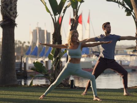 A couple practices yoga on a grassy waterfront lawn with palm trees, stretching in unison near sailboats.