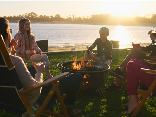 Friends gather around a fire pit on a lakeside lawn at sunset, chatting and toasting drinks.