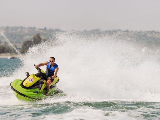 A person on a bright green jet ski speeds across open water, blasting a huge splash into the air.