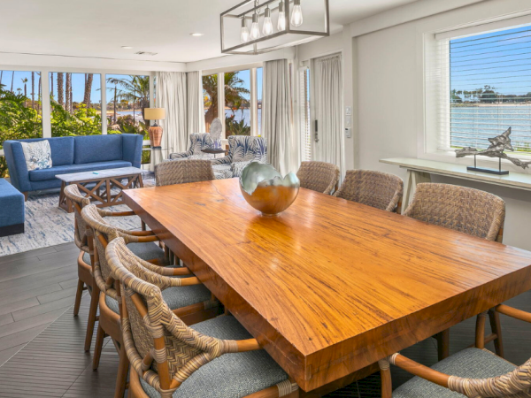 A bright modern living and dining room with a large wooden table, rattan chairs, plush seating, and windows facing the water.