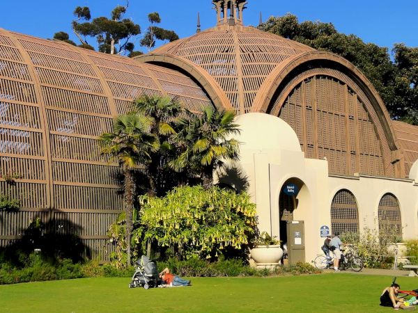 A sunny garden in front of a large dome with wooden lattice and arches, with palm trees and people relaxing.