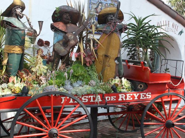 A red cart planter labeled &ldquo;ASTA DE REYES&rdquo; holds succulents, with three metal soldier-like statues in a garden display.