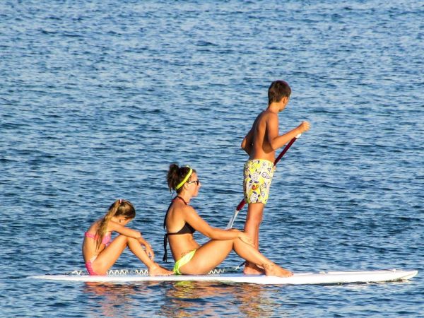 A family paddles on a stand-up board in the ocean as one child splashes, with another sitting nearby.