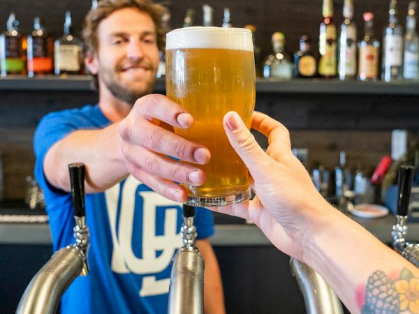 A bartender and customer clink a pint of amber beer in a pub with taps and bottles in the background.