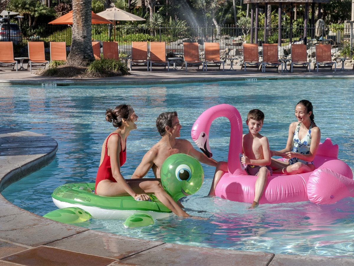 Kids splash in a sunny pool, riding colorful inflatable floats near lounge chairs.