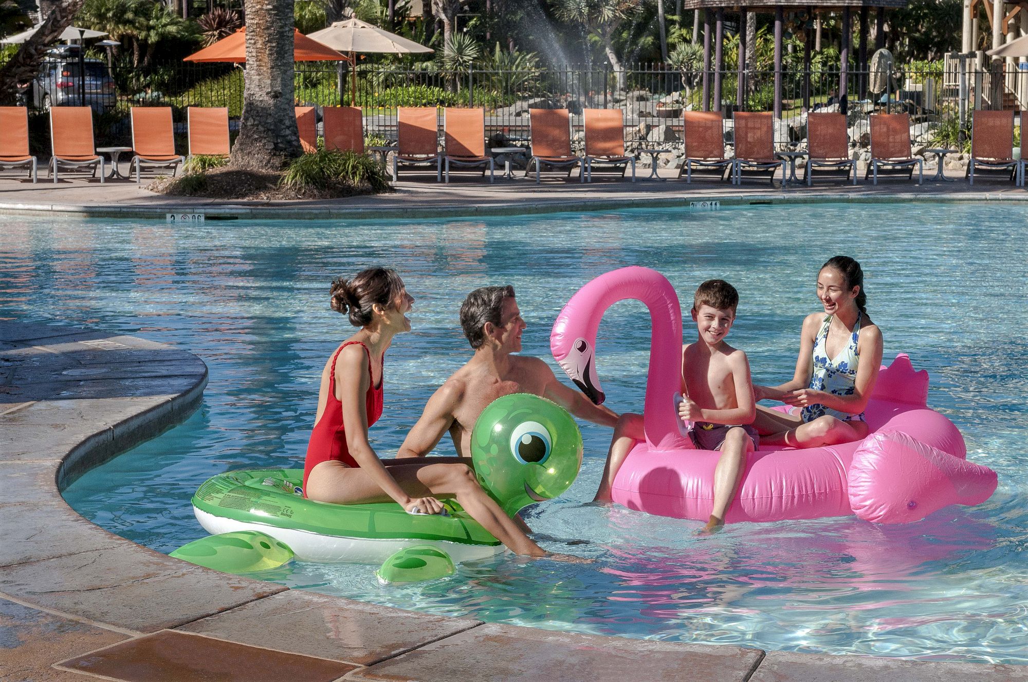 Kids play in a sunny hotel pool on colorful inflatable floaties, with lounge chairs and trees in the background.