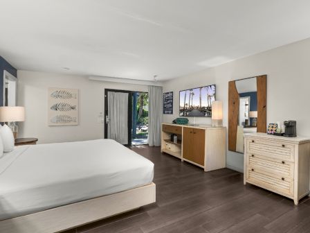 A bright bedroom with white bed, dresser, wall art, coffee maker, and a sliding door to a sunny patio.