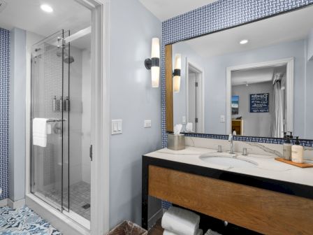 A modern bathroom with a glass shower, marble vanity and mirror, patterned blue tile walls, and bright lighting.