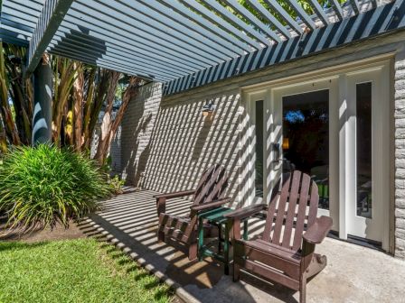 Sunlit outdoor patio with two rocking Adirondack chairs under a striped pergola beside a brick wall and lush plants.