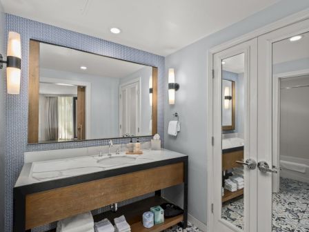 Modern hotel bathroom with a double sink vanity, mirrored walls, wall lights, patterned tile floors, and neatly folded towels.
