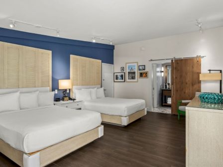 A modern hotel room with two white beds, blue accent wall, framed photos, a dresser, and a wooden door with an open doorway.
