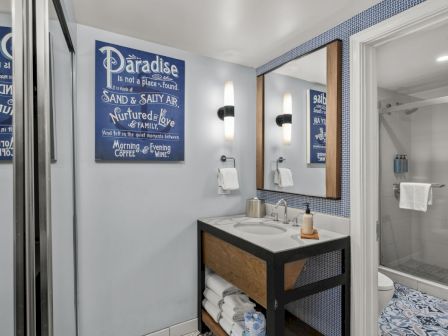 A modern hotel bathroom with double vanity sinks, towel rails, shower glass, blue patterned tile, and framed wall art reading &ldquo;Paradise&rdquo;.