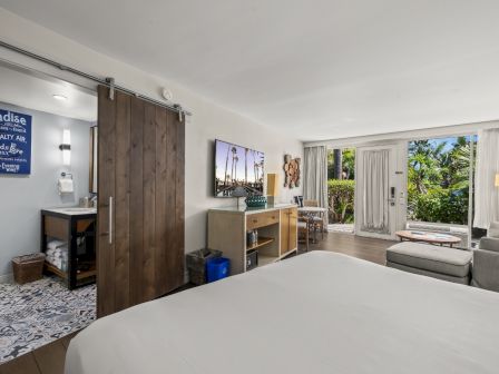 A modern hotel bedroom with a bed, vanity/dresser, sliding wooden door, and big windows overlooking green palm scenery.