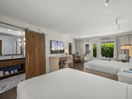 A modern hotel room with a large bed, wood doors, a vanity mirror, and outdoor-view windows with curtains, in bright daylight.