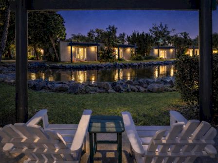 A nighttime backyard patio with white chairs overlooking a lit pond and garden, with a house glowing in the distance.