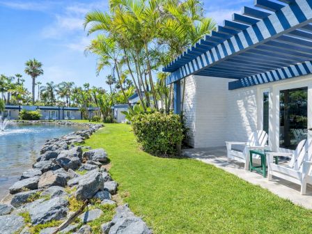 A tropical resort scene with a pond-like pool, palm trees, and white patio chairs under a blue-striped awning.