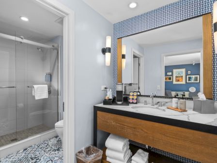Modern hotel bathroom with a marble sink vanity, round lights, patterned wall, and a glass shower with towels.