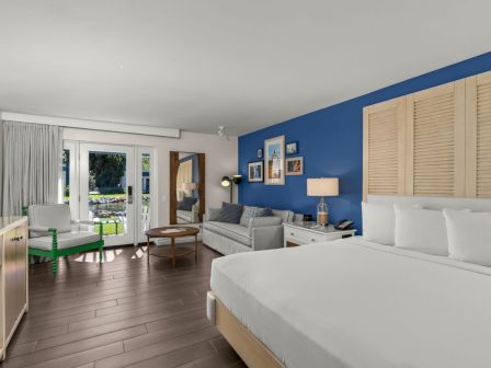 A bright blue bedroom with a king bed, sofa, framed art, and large glass doors opening to a garden.