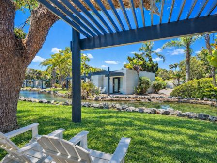 A tropical resort garden with a blue pergola over a lush lawn, stones by a small pool, and modern villa walls.