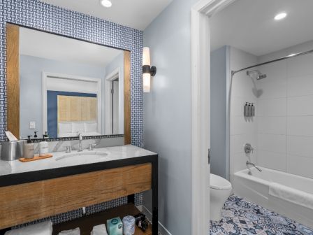 A modern hotel bathroom with a wooden vanity, large mirror, and a shower-tub combo with patterned floor tiles.