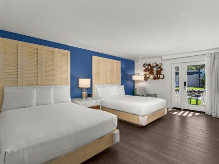 A bright bedroom with two white beds, blue accent wall, lamps, and open doors to a green patio.