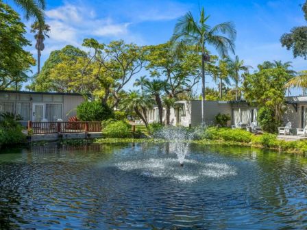 A tropical resort scene with a pond fountain, palm trees, lush greenery, and bungalow cabins under a bright blue sky.
