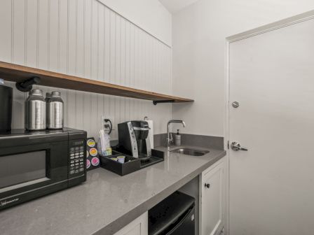A small kitchenette with a microwave, coffee maker, stacked coffee supplies, and a stainless sink against white cabinets.