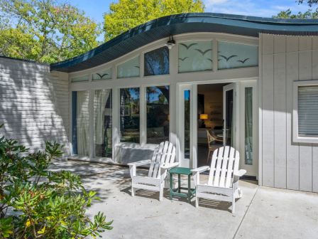 A bright backyard patio beside a white house, with white rocking chairs, open doors/windows, and lush green shrubs and trees behind.