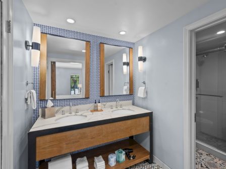 A modern bathroom with double sinks on a wood vanity, blue patterned tile mirrors, and a glass-enclosed shower.