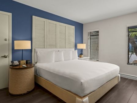 A bright bedroom with a white bed, two lamps, a navy accent wall, and windows with blinds.