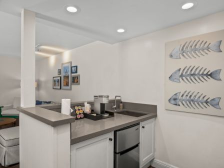 A modern kitchenette with a sink and coffee maker, white cabinets, and wall d&eacute;cor featuring three metal fish on a beige panel.