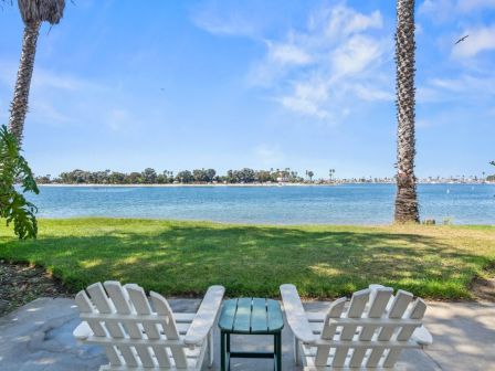White Adirondack chairs on a patio overlook a calm blue ocean, palm trees framing the view under a sunny sky.