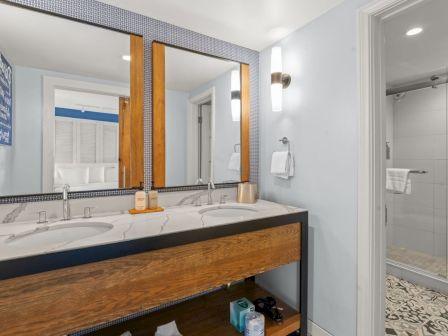 A modern hotel bathroom with a marble double-sink vanity, large mirrors, toiletries, and a glass shower with folded towels.