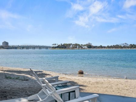 A seaside view with white lounge chairs, calm blue water, and a distant pier/bridge under a bright sky.