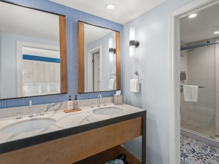 A modern bathroom with a double vanity, framed mirrors, wall lights, towels, and a glass shower with tiled floor and patterned rug.
