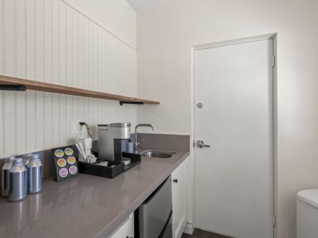 A small kitchenette with coffee maker, cans and cups, a sink, and a nearby toilet beside a closed door.
