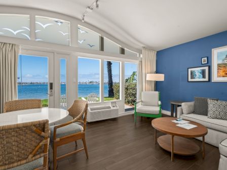 A bright seaside living room with beige sofas, woven chairs, and large windows showing the ocean under a blue sky.