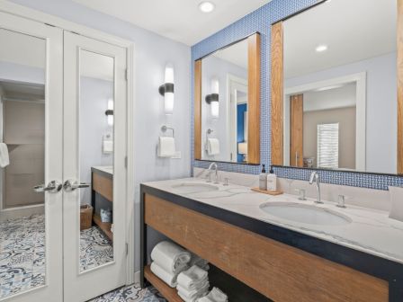 A modern bathroom with double sinks, a blue-tiled wall, wooden cabinetry, patterned floor, and mirrored vanity lighting.