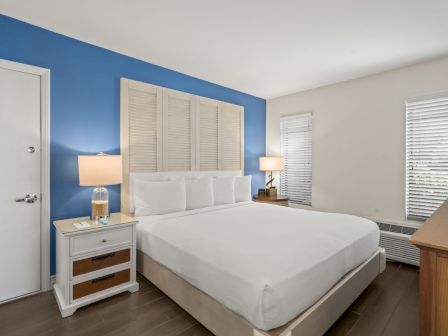 A bright bedroom with a white bed, blue accent wall, bedside lamps, shutters, and a neatly made room with wooden furniture.