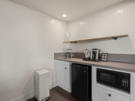A small kitchenette with sink, coffee machine, microwave, white cabinets, and a trash bin in a bright room with paneled walls.
