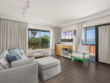 A modern living room with a gray sofa, windows and curtains, and a view of palm trees and the ocean beyond the balcony door.