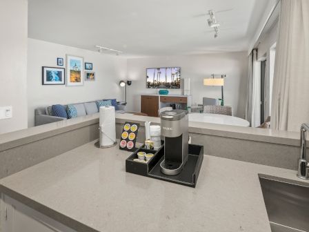 Modern living room with a sofa and artwork, plus a counter/kitchenette featuring a Keurig coffee machine, with patio doors.