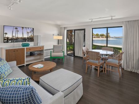 A bright modern living room with a sofa, coffee table, TV, dining set, and floor-to-ceiling windows overlooking the ocean.