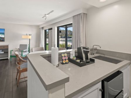 A modern room with a white kitchen island, coffee machine and sink, plus chairs and curtains with a view outside.