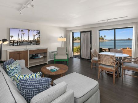 Bright living room with a gray sofa, TV, dining set, and open doors showing an oceanfront view.