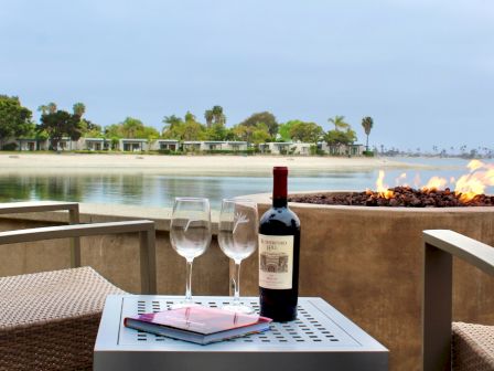 A beachside patio with two wine glasses, a bottle, and a firepit overlooking a calm ocean shoreline.