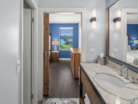 A modern bathroom with double sinks, a mirror, wall lights, a wooden vanity, and a view into a blue bedroom.