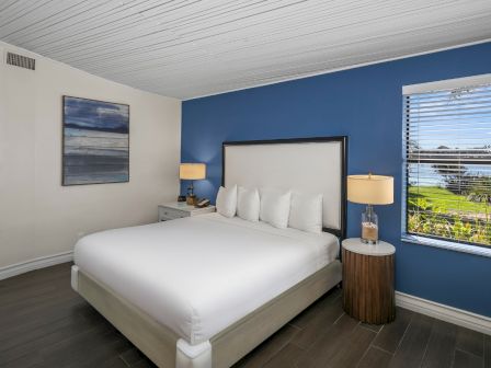 A modern bedroom with a white bed, blue accent wall, bedside lamps, and a window with blinds.