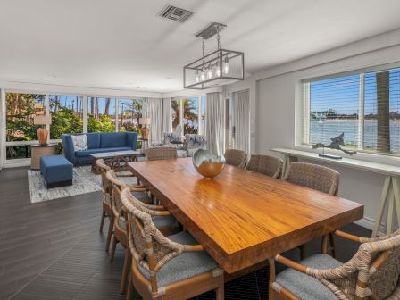 A sunlit modern dining room with a large wooden table, wicker chairs, and big windows overlooking a waterfront view.